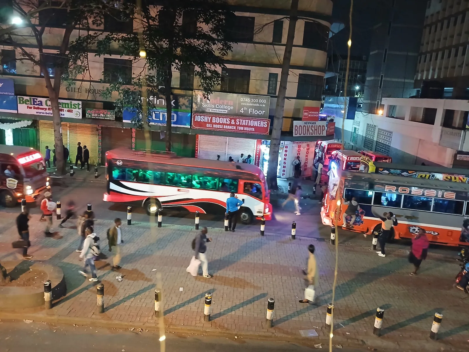 Pedestrians walking past a bus and decorated matatus on Tom Mboya Street at night